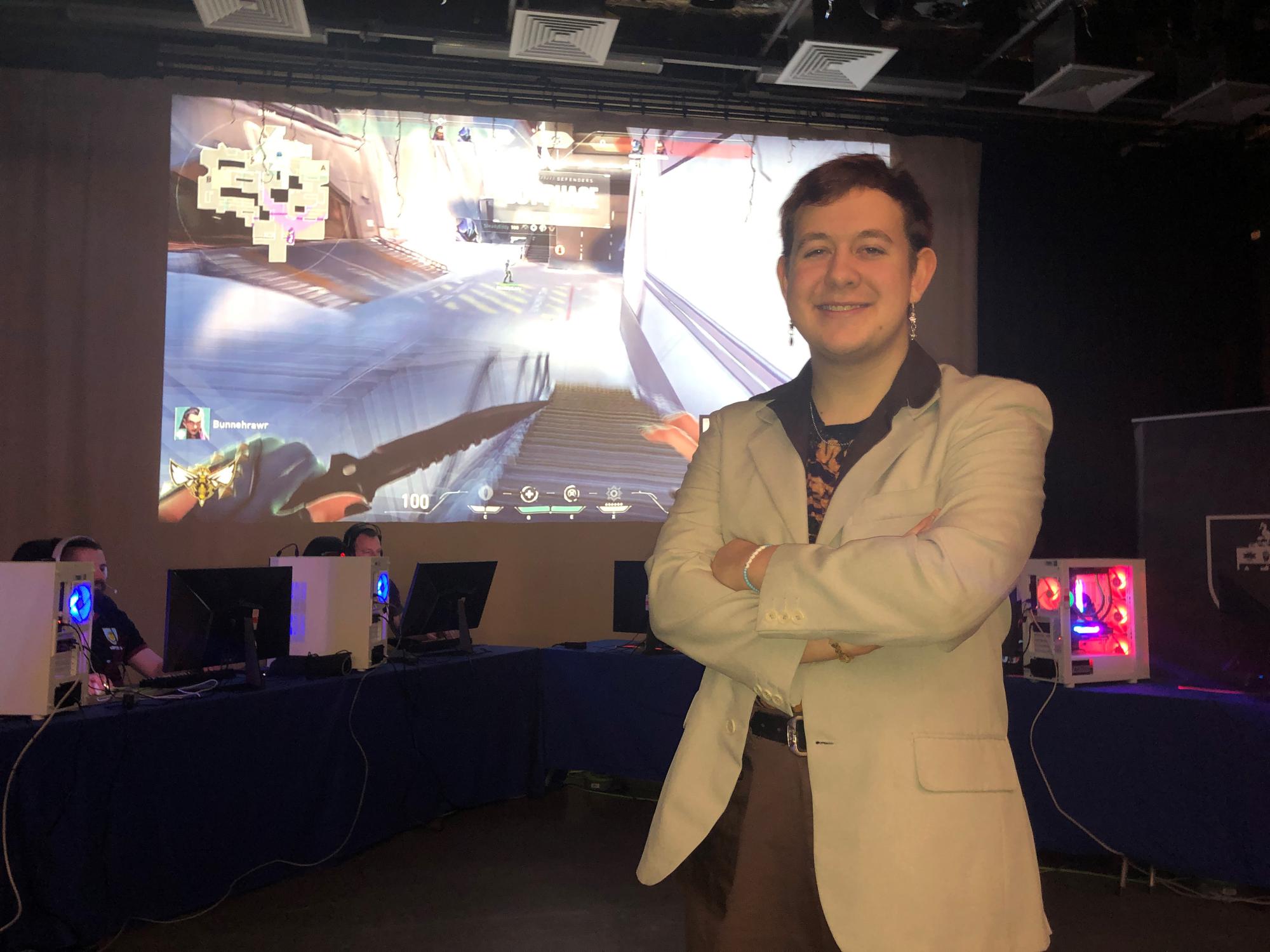 Student sands proud before a giant screen as his colleagues and peers play Valoran in a charity tournament behind him